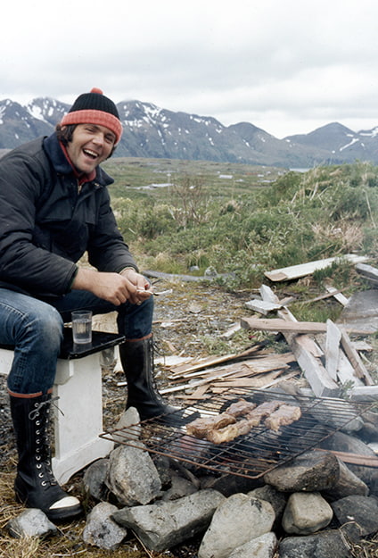 Dr. James Estes sitting on a bench and cooking meat over a fire. A mountain range is behind him.