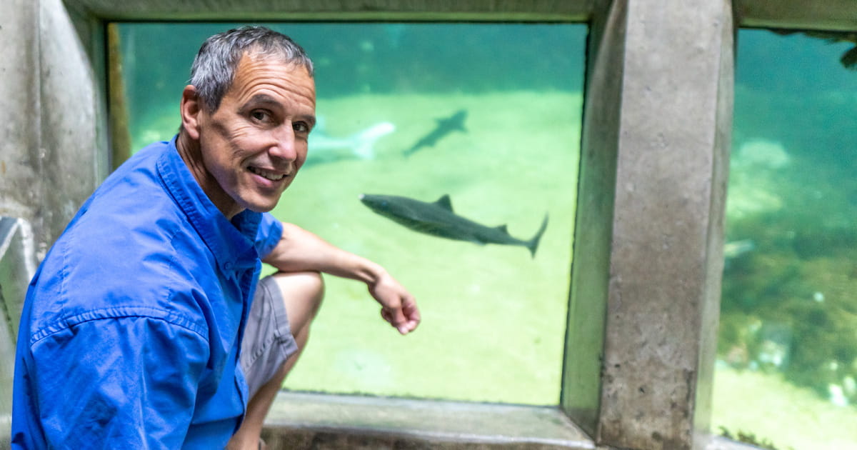 Chris Van Damme kneeling in front of a large viewing window into the Underwater Dome at the Seattle Aquarium as a dogfish swims inside the dome behind him.