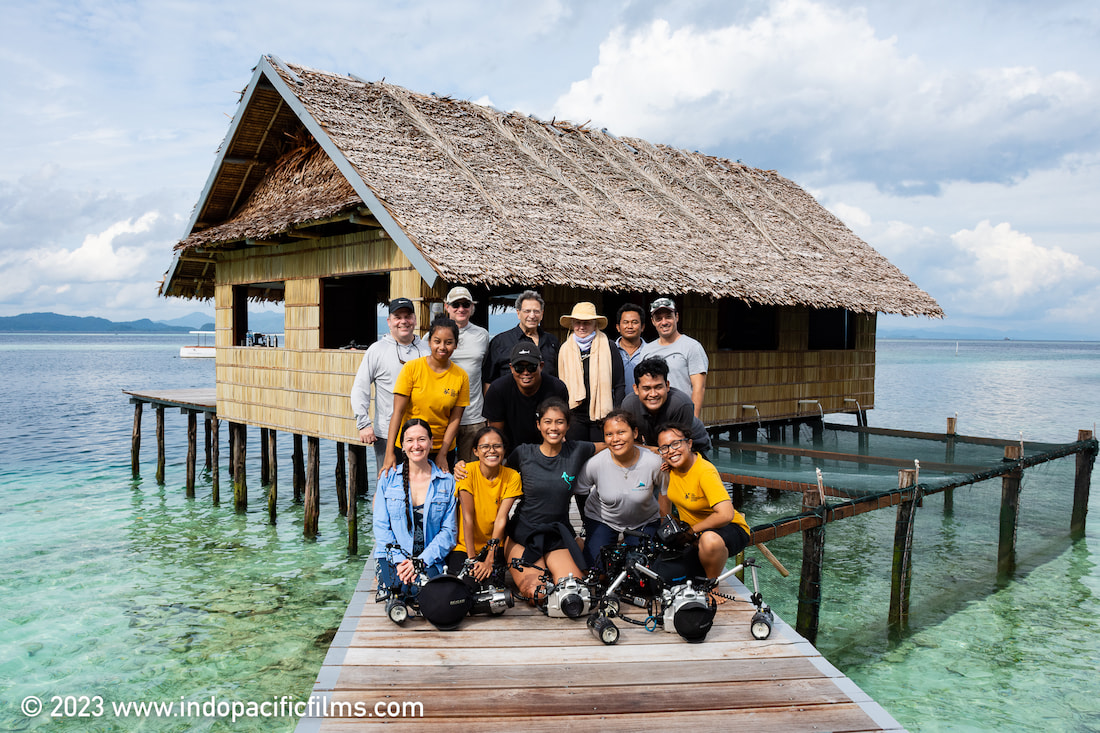 Members of the ReShark collective pose on a wooden walkway for a group photo in front a hut built over shallow ocean waters in Indonesia.