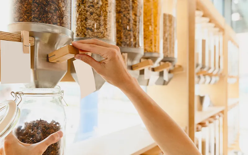 A person refilling a reusable glass jar with granola from a bulk item area inside a store.