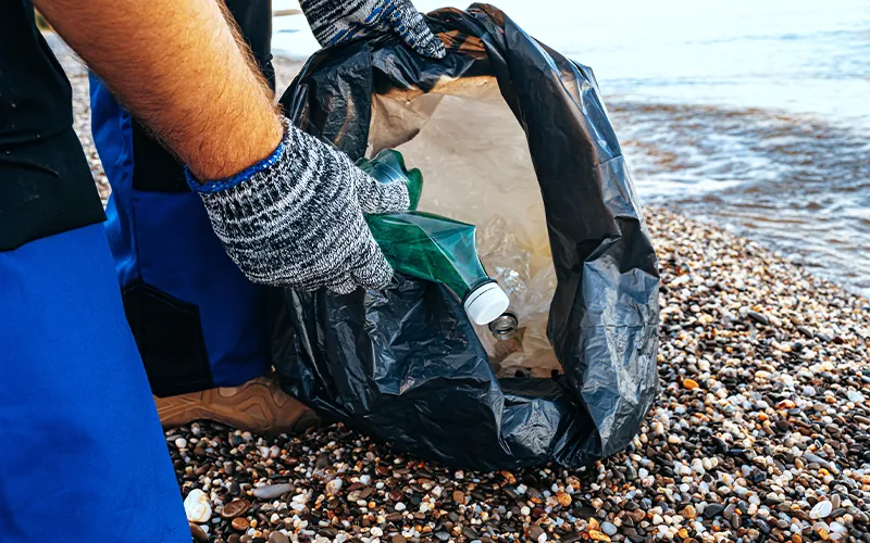 An individual kneeling down on a rocky beach, picking up a plastic bottle and placing it inside a garbage bag which has other items already inside.