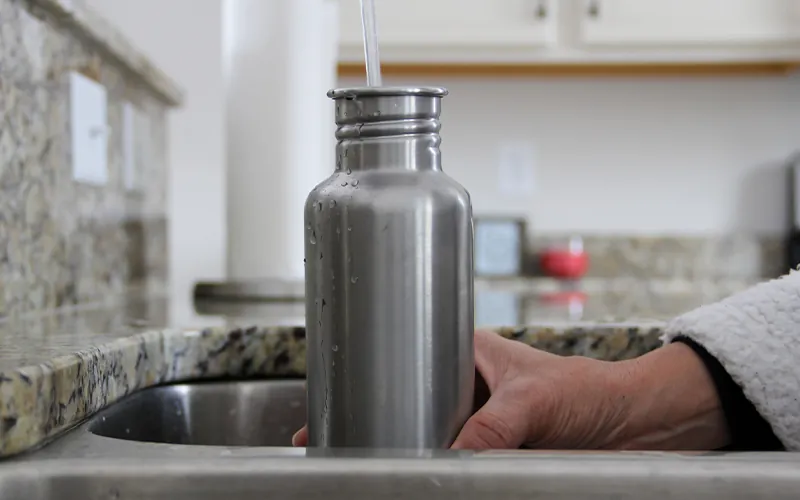 A person holding a reusable water bottle under the water tap of a sink, filling the bottle.