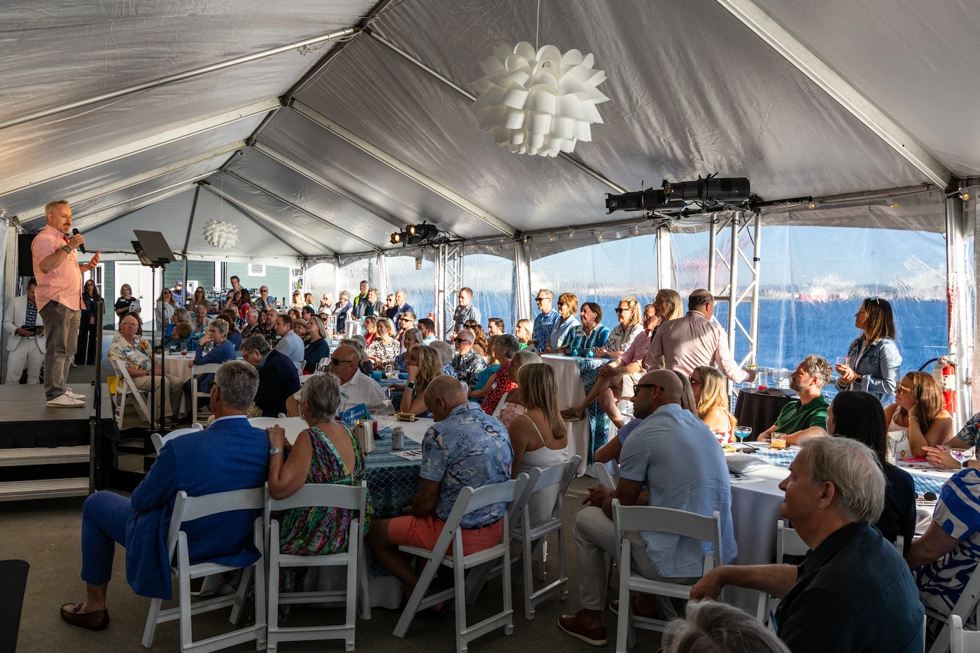 Guests at the Seattle Aquarium’s annual Splash event gathered under a tent with transparent sides, allowing for a view of a body of water and a sunset in the background. Guests listen to Jim Wharton speak on a stage at the front as he speaks into a microphone while facing the seated audience.