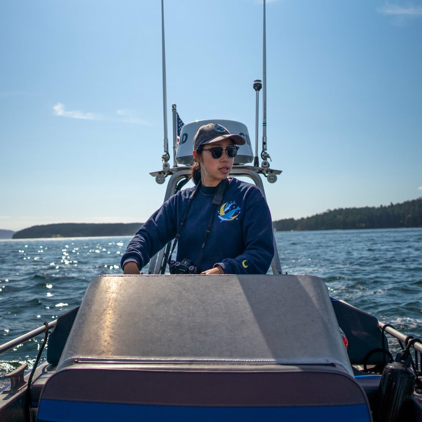 Bethany Shimasaki driving a boat on the open water. She is wearing a blue sweatshirt, a grey baseball cap, and sunglasses. An outcropping of forested islands can be seen in the far distance.