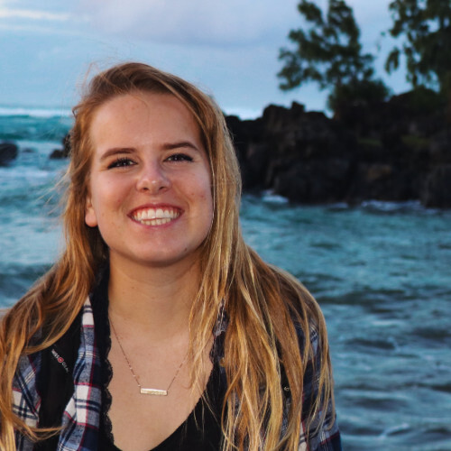 A photo of Emma Strand standing in front of the ocean. She has long, straight blonde hair.