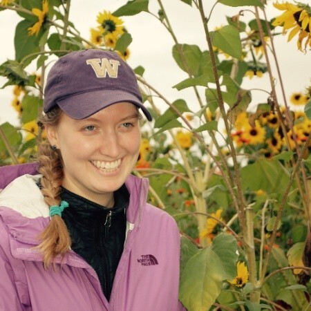 Liz Allyn standing in front of a sunflower patch. She is wearing a pink Northface jacket and a purple University of Washington baseball cap.