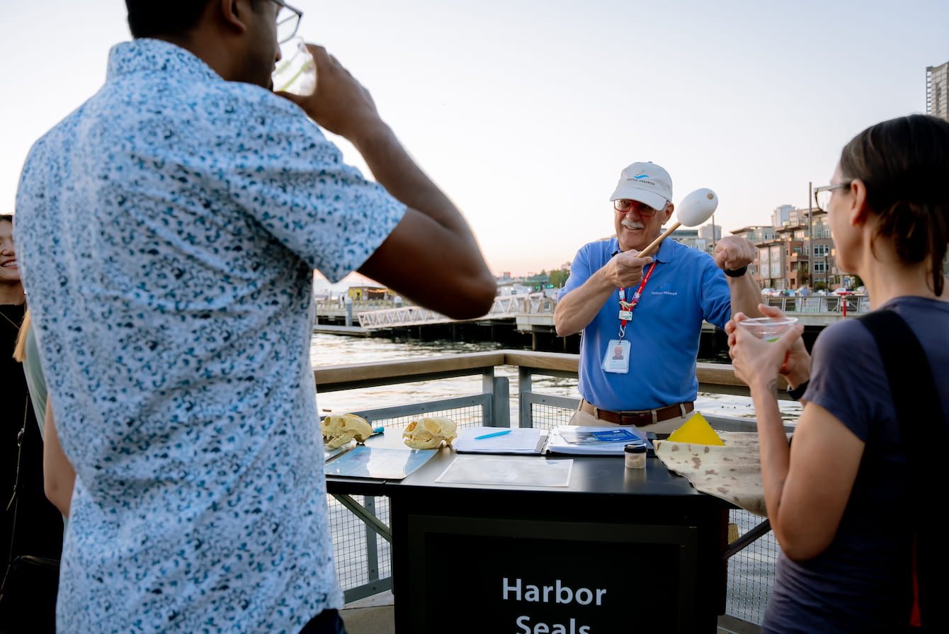 An Aquarium volunteer giving a demonstration to two guests holding drinks.