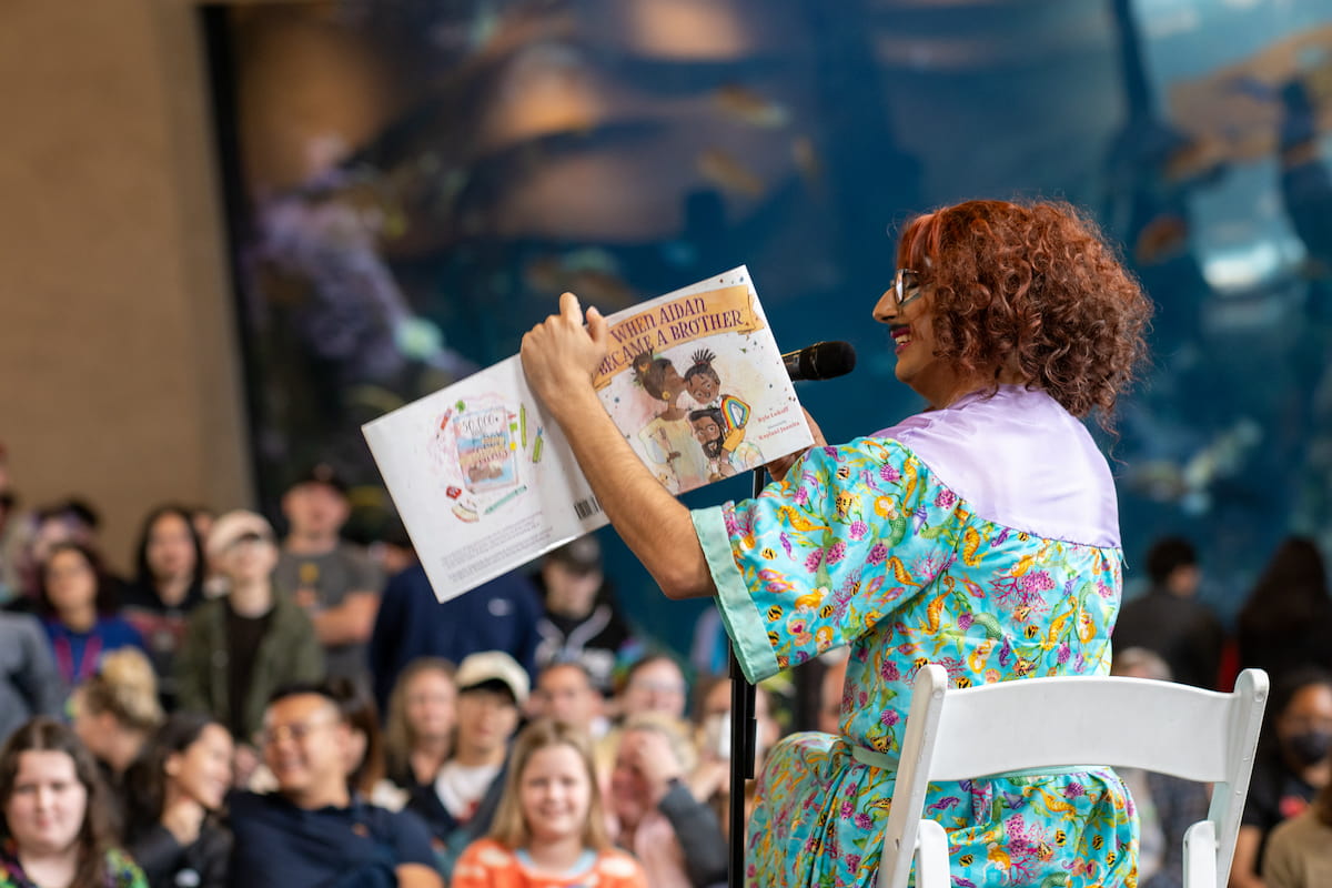 A drag queen in a blue dress reading a children's book to a crowd of people.