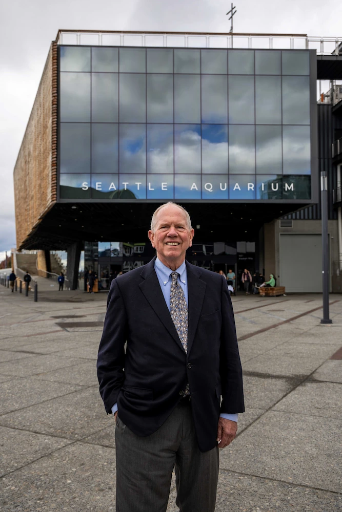 Bob Davidson stands, smiling, in front of the Seattle Aquarium's Ocean Pavilion building. Large glass windows on the upper front of the building reflect a partly cloudy sky. The word Seattle Aquarium are displayed across the bottom of the windows.