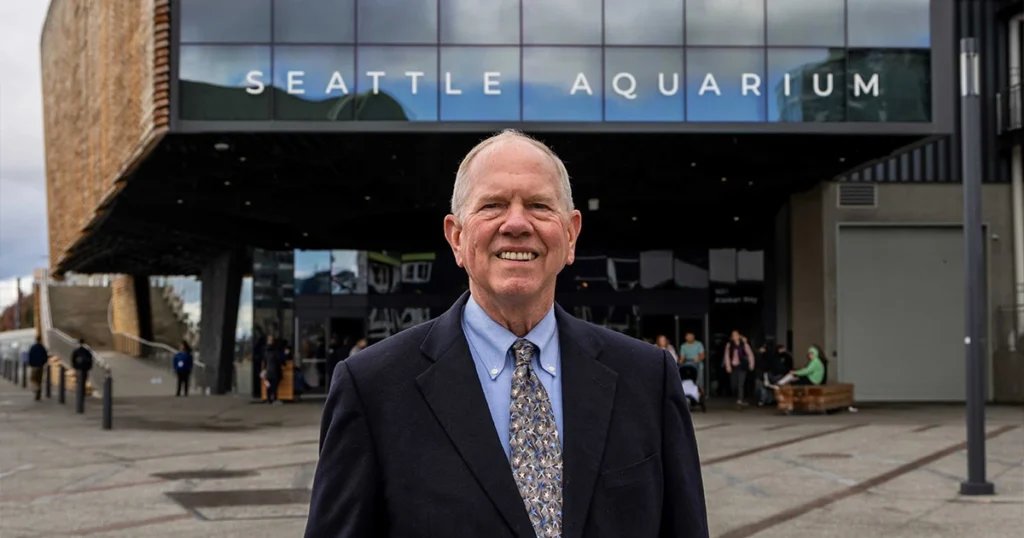 Bob Davidson stands, smiling, in front of the Seattle Aquarium's Ocean Pavilion building. Large glass windows on the upper front of the building reflect a partly cloudy sky. The word Seattle Aquarium are displayed across the bottom of the windows.