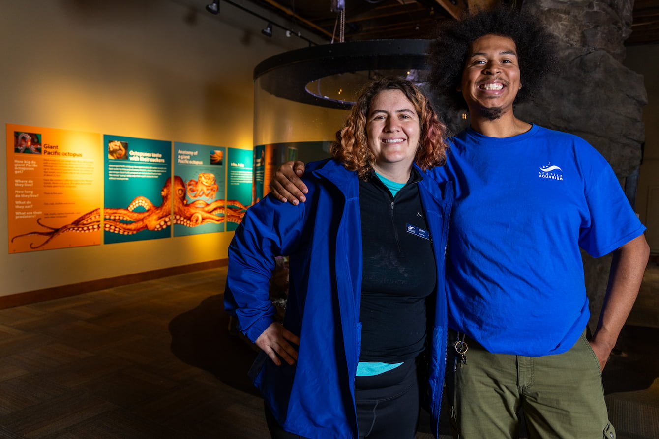 Aquarists Malik Johnson and Kaitlin Brawley standing in front of the Aquarium's octopus habitat with their arms around each other's shoulders.