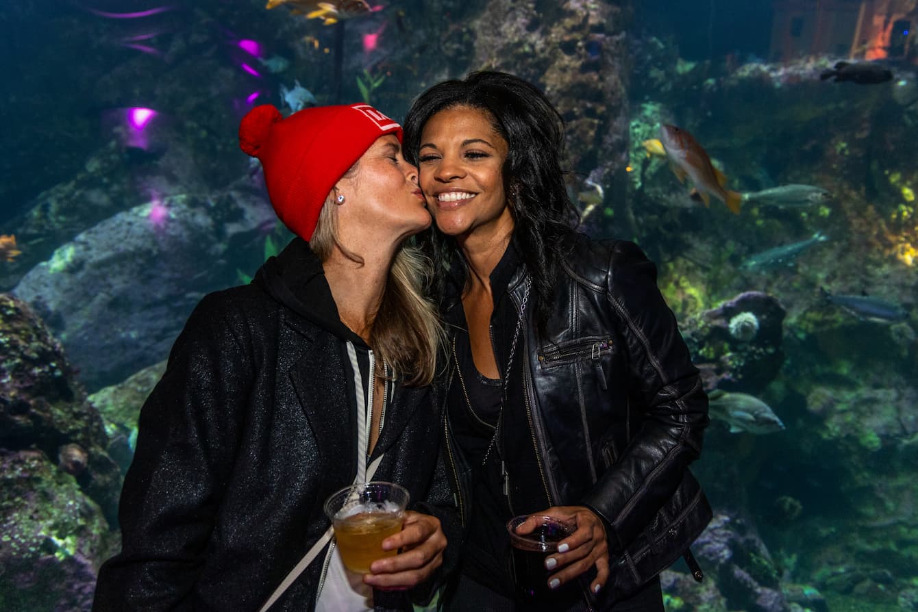 Two people holding drinks and standing in front of the Window on Washington Waters habitat. One person is kissing the other on the cheek.