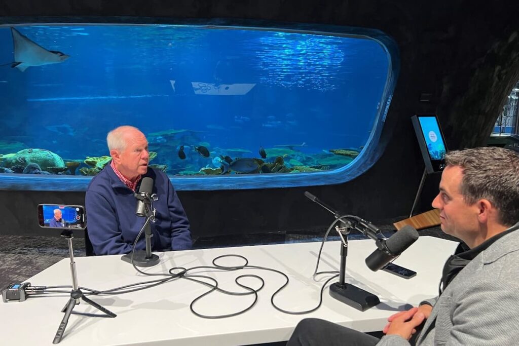 Jon Scholes and Bob Davidson sitting at a table with microphones in front of The Reef habitat in the Ocean Pavilion.