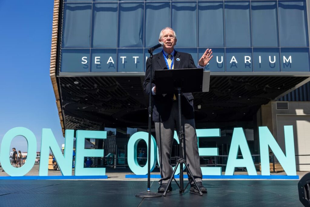 Bob Davison speaking into a microphone in front of the Seattle Aquarium's Ocean Pavilion.