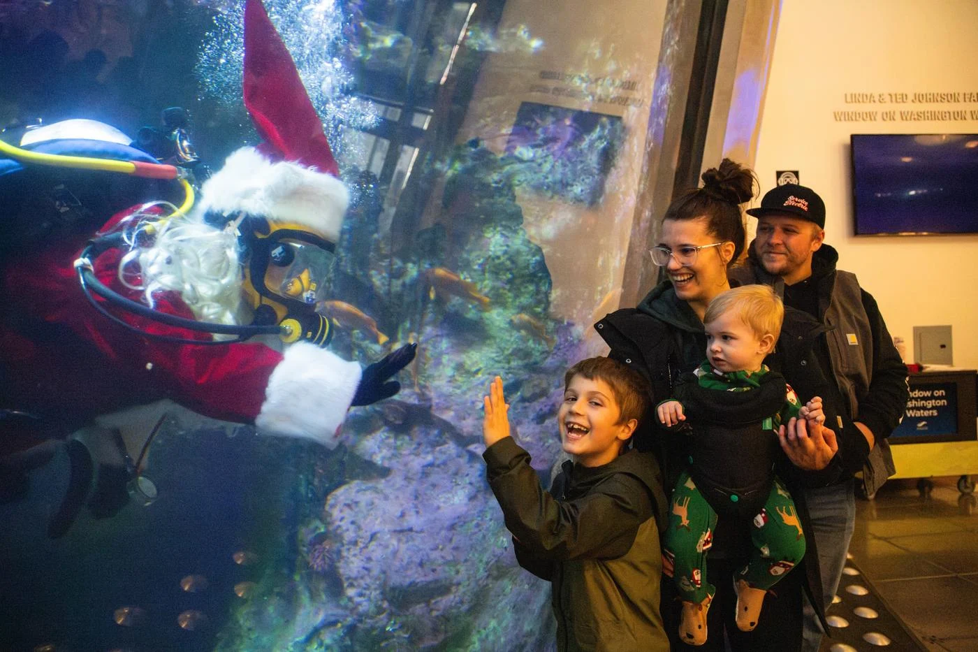 A young boy and his family pose for a photo against the large glass viewing window of the Seattle Aquarium Window on Washington Water habitat alongside a scuba diver dressed as Santa swimming inside the habitat.