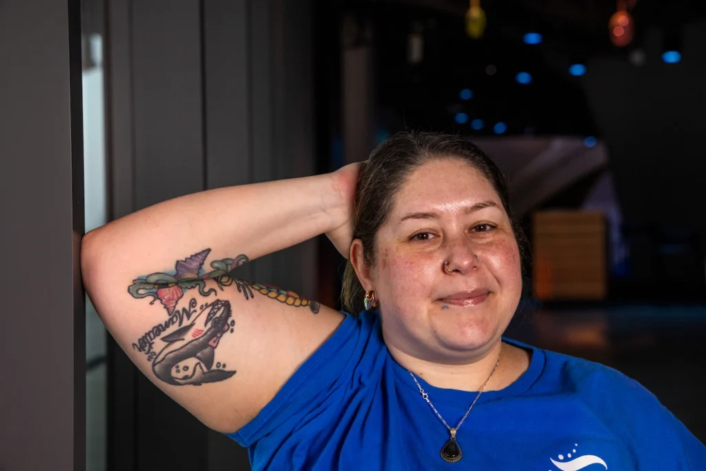 Amber, a woman with dark hair tied back in a ponytail, holding her right arm up to showcase a shark tattoo on her bicep.