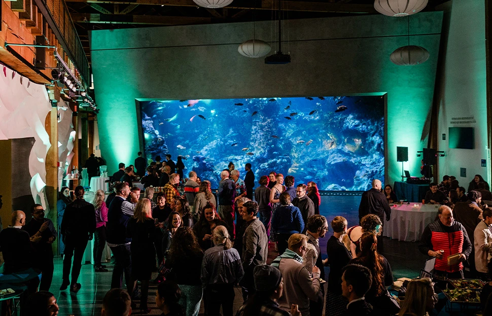 A large group of adults attending a private event hosted in the Seattle Aquarium's Puget Sound Hall, against a backdrop of the large viewing window into the Window on Washington Waters habitat.