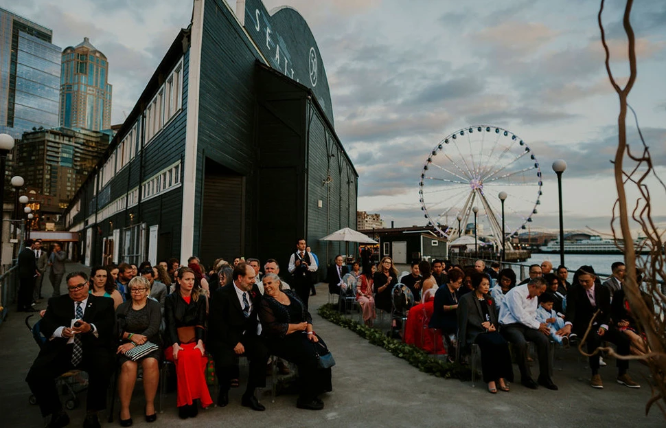 Wedding guests in formal attire sitting in chairs for a wedding ceremony outside the back of the Seattle Aquarium's Pier 59 building. The Seattle Great Wheel and a ferry are to the right of the guests as they wait for the ceremony to begin.