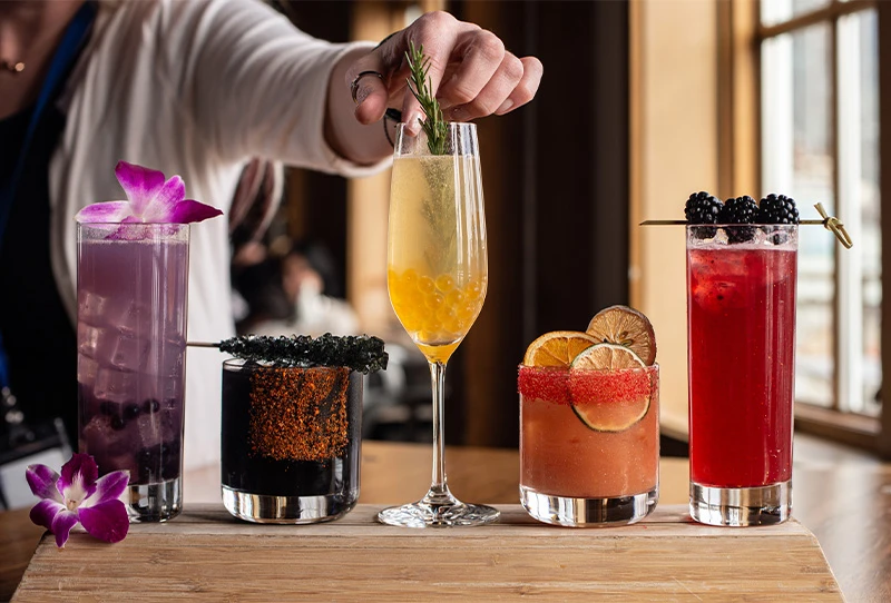 Five different multicolored mixed drinks lined up on a table top. Each drink is in a different shaped glass with unique garnishes including flower petals, rock candy, limes and blackberries.